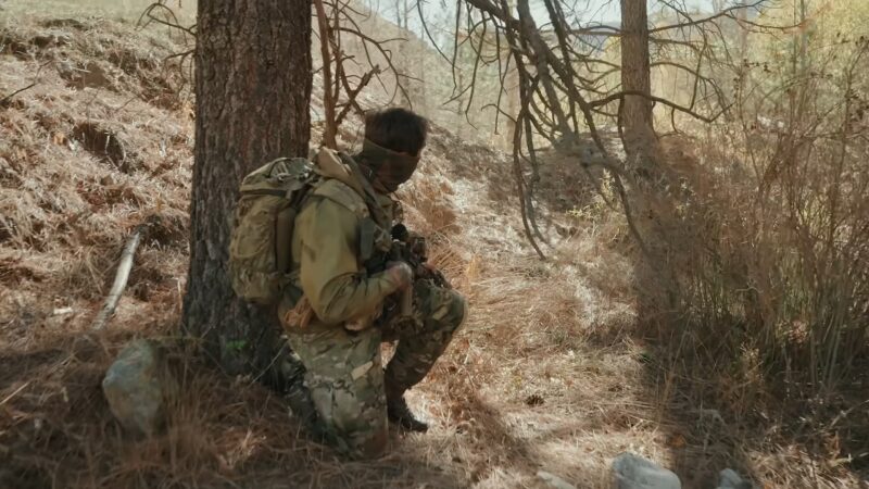 A Soldier in Camouflage Gear Crouches by A Tree in A Wooded Area, Aiming a Rifle During a Tactical Military Training Exercise