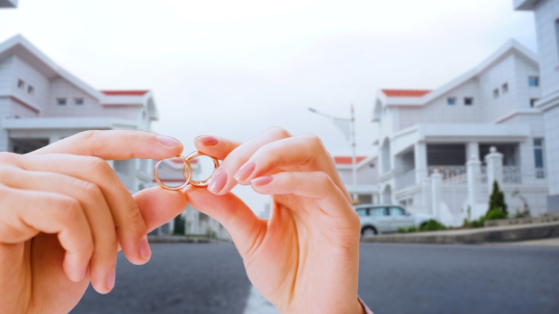 Hands Holding Wedding Rings in Front of A Residential Area, Representing how Marital Status Influences Military Housing Support