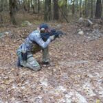 Person Wearing Military Fashion, Including Camouflage and Tactical Gear, Takes a Kneeling Position While Aiming at A Gun Range