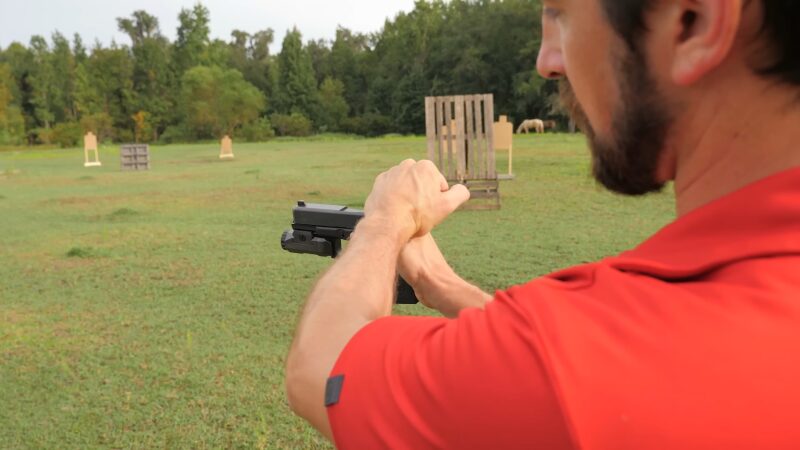 A Man in A Red Shirt Is Carefully Aiming a Handgun at A Target in An Open Field, Staying Aware of His Surroundings