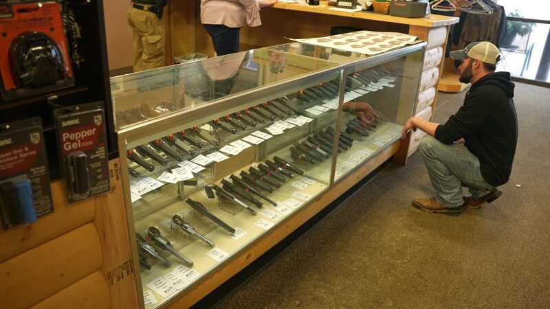 A Man Is Crouched Down, Carefully Looking at A Selection of Handguns in A Glass Display Case at A Gun Store