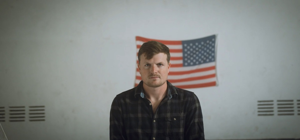 Man in casual clothing seated with a U.S. flag in the background