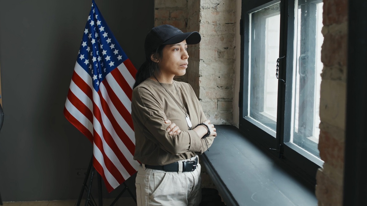 Woman in military-style attire standing by a window with the U.S. flag in the background