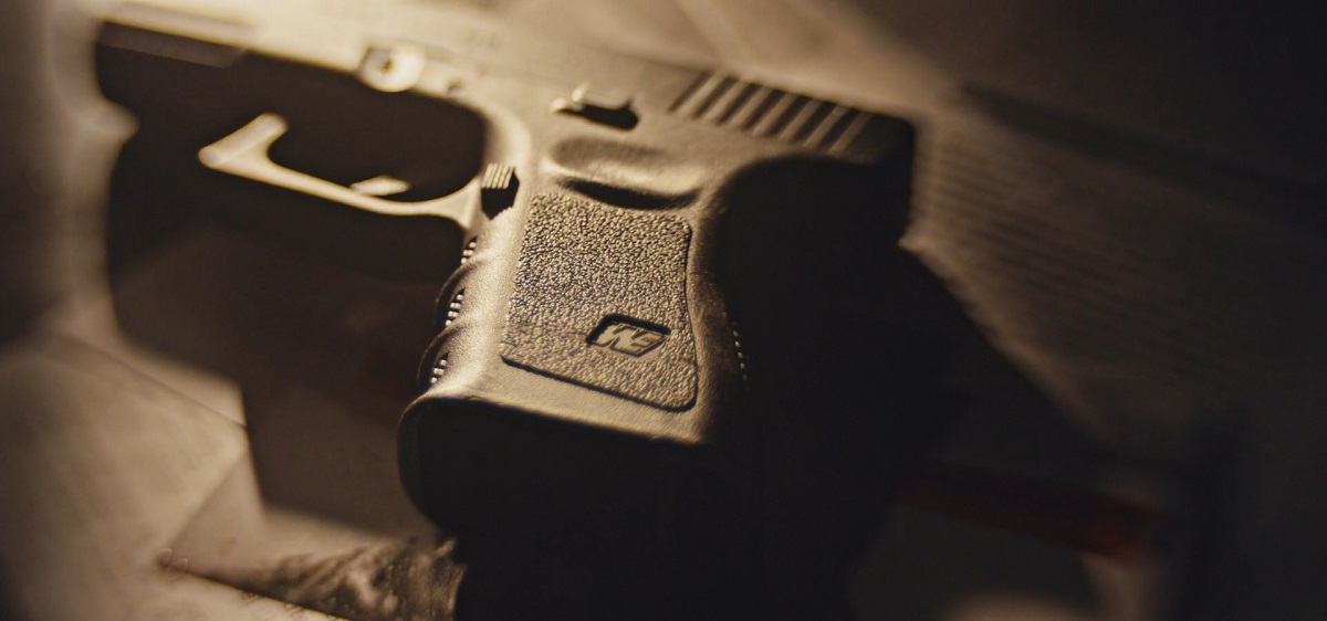 A close-up of a black handgun placed on a surface with dramatic lighting casting shadows on the textured grip