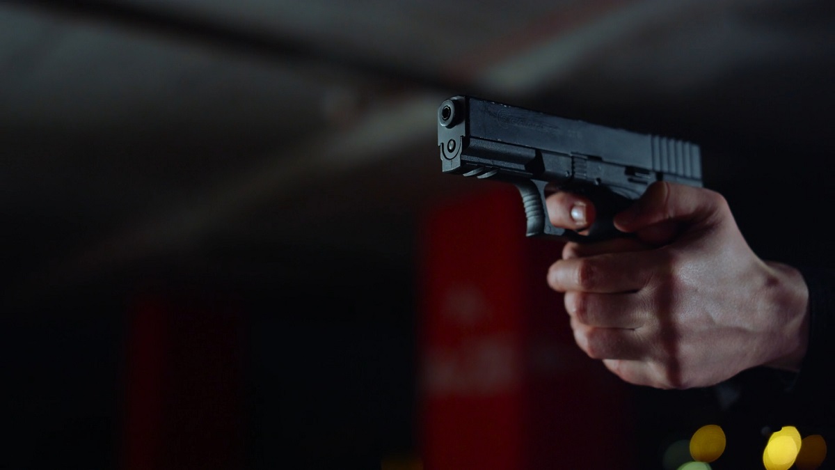 A person aiming a black semi-automatic handgun in a dimly lit indoor setting, with a red structural column in the background