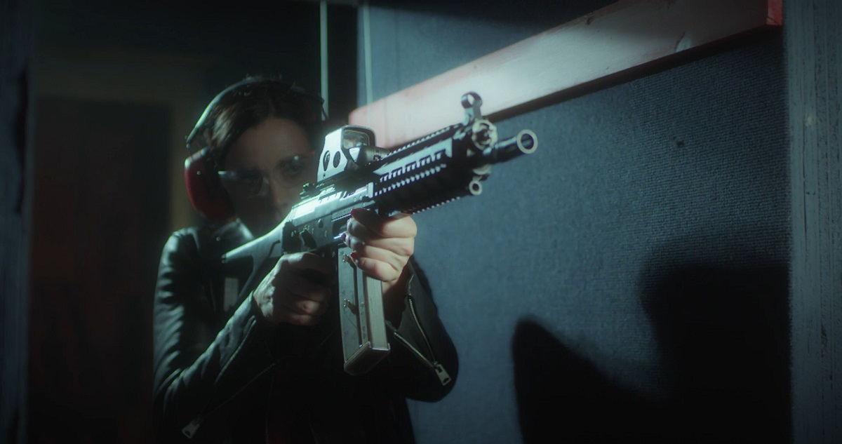 A woman at an indoor shooting range aiming a tactical rifle with a scope, wearing safety glasses and ear protection