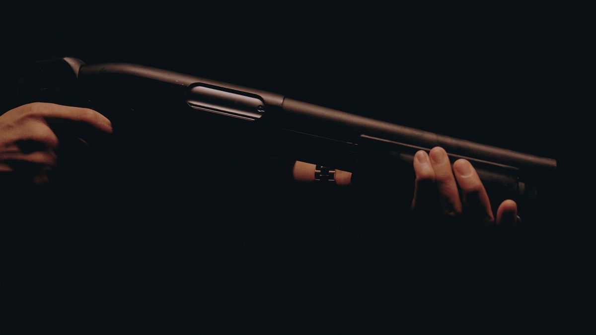 A close-up shot of a person holding a black shotgun in low light, with hands gripping the barrel and pump-action mechanism